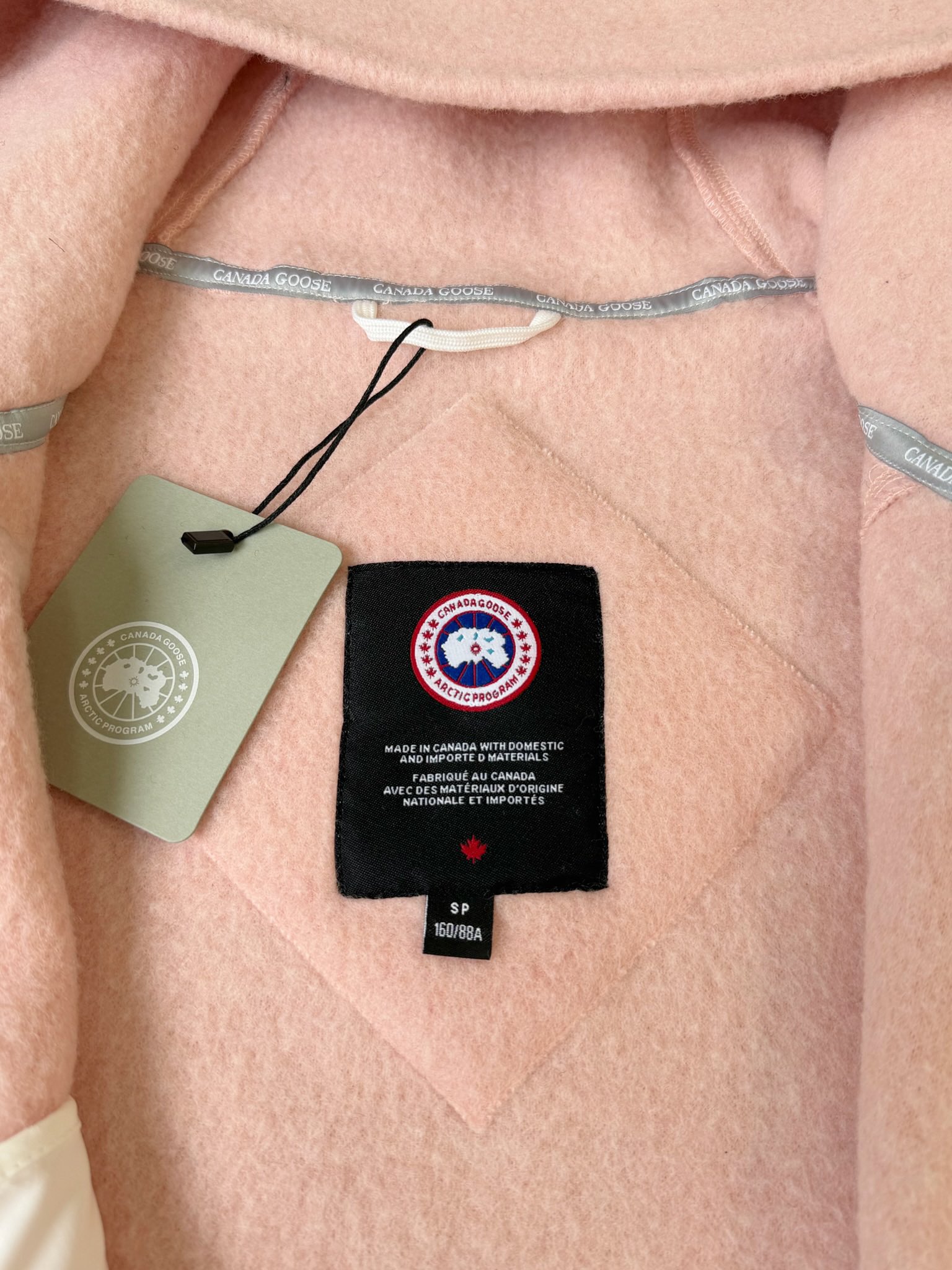 大鹅/Canada Goose抓绒羊毛饰章连帽夹克外套 面料选择: 以羊毛为主体，加入极少量氨纶和聚酯纤维混纺而成的羊毛抓绒面料。羊毛作为核心天然成分，提供无与伦比的温度调节、抑菌防臭和亲肤感。少量氨纶的喂入赋予了面料一定的四向弹性，日常活动时毫无束缚感。作为抓绒结构的基础，聚酯纤维增强了面料的耐久性、抗皱性和快干性，它构成了抓绒的蓬松感，并提供锁住空气等功能。 细节1: 衣袖和下摆采用2乘2罗纹收口，其强大的弹性，可以紧密贴合不同粗细的手腕和腰部，形成一个有效的&lsquo;密封&rsquo;,防止冷风倒灌。在多次拉伸和使用后仍能保持原有形状，不易松垮变形。且罗纹袖口能传递一种设计感，强化视觉美观度。 细节2: 兜帽设计与松紧扣的搭配提升穿戴时的保暖度与防风性能。 辅料:定制弹性抽绳松紧扣定制拉链吊坠定制臂章 尺码：S M L 颜色：黑 灰 咖 米 粉