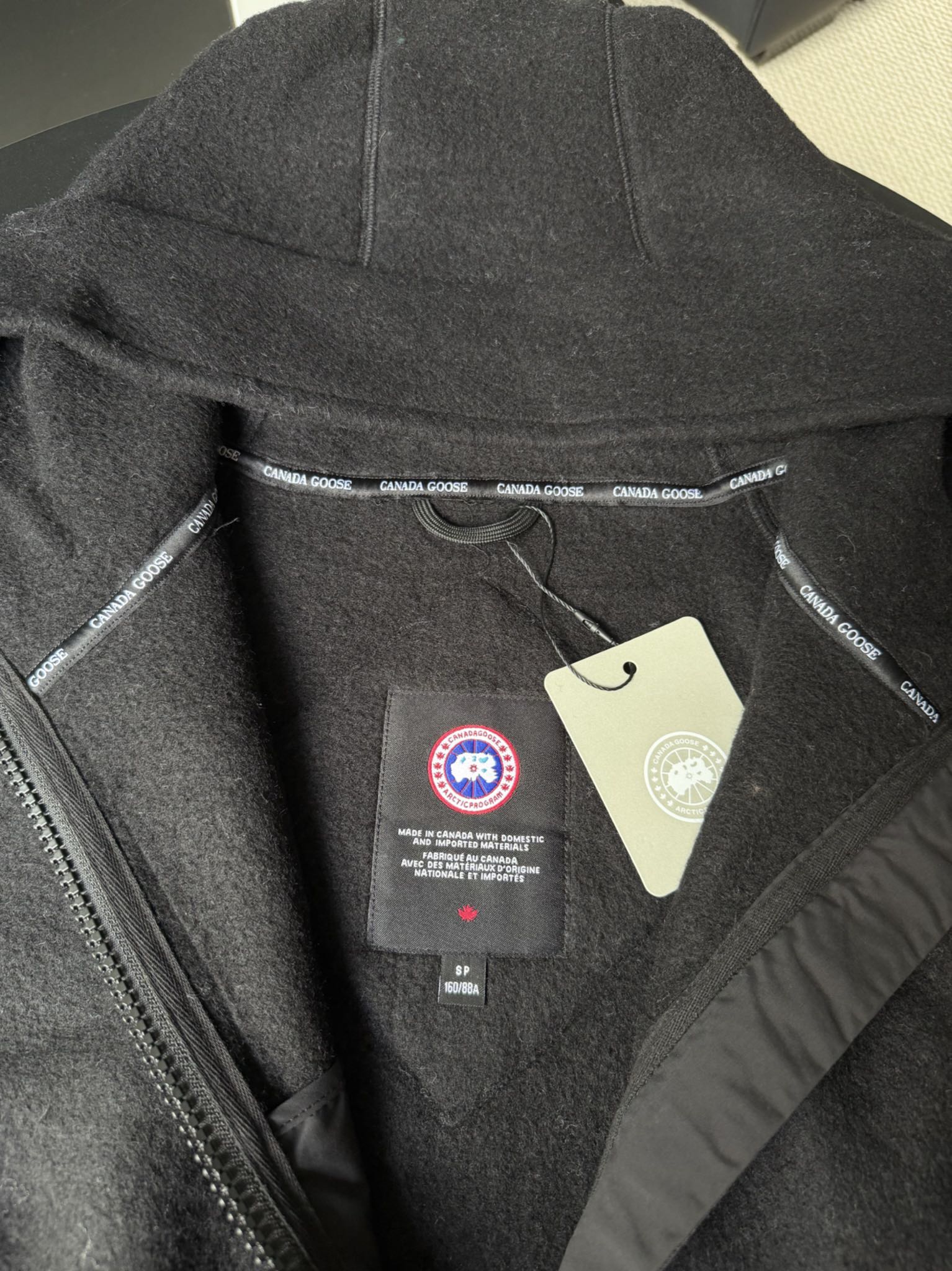 大鹅/Canada Goose抓绒羊毛饰章连帽夹克外套 面料选择: 以羊毛为主体，加入极少量氨纶和聚酯纤维混纺而成的羊毛抓绒面料。羊毛作为核心天然成分，提供无与伦比的温度调节、抑菌防臭和亲肤感。少量氨纶的喂入赋予了面料一定的四向弹性，日常活动时毫无束缚感。作为抓绒结构的基础，聚酯纤维增强了面料的耐久性、抗皱性和快干性，它构成了抓绒的蓬松感，并提供锁住空气等功能。 细节1: 衣袖和下摆采用2乘2罗纹收口，其强大的弹性，可以紧密贴合不同粗细的手腕和腰部，形成一个有效的&lsquo;密封&rsquo;,防止冷风倒灌。在多次拉伸和使用后仍能保持原有形状，不易松垮变形。且罗纹袖口能传递一种设计感，强化视觉美观度。 细节2: 兜帽设计与松紧扣的搭配提升穿戴时的保暖度与防风性能。 辅料:定制弹性抽绳松紧扣定制拉链吊坠定制臂章 尺码：S M L 颜色：黑 灰 咖 米 粉