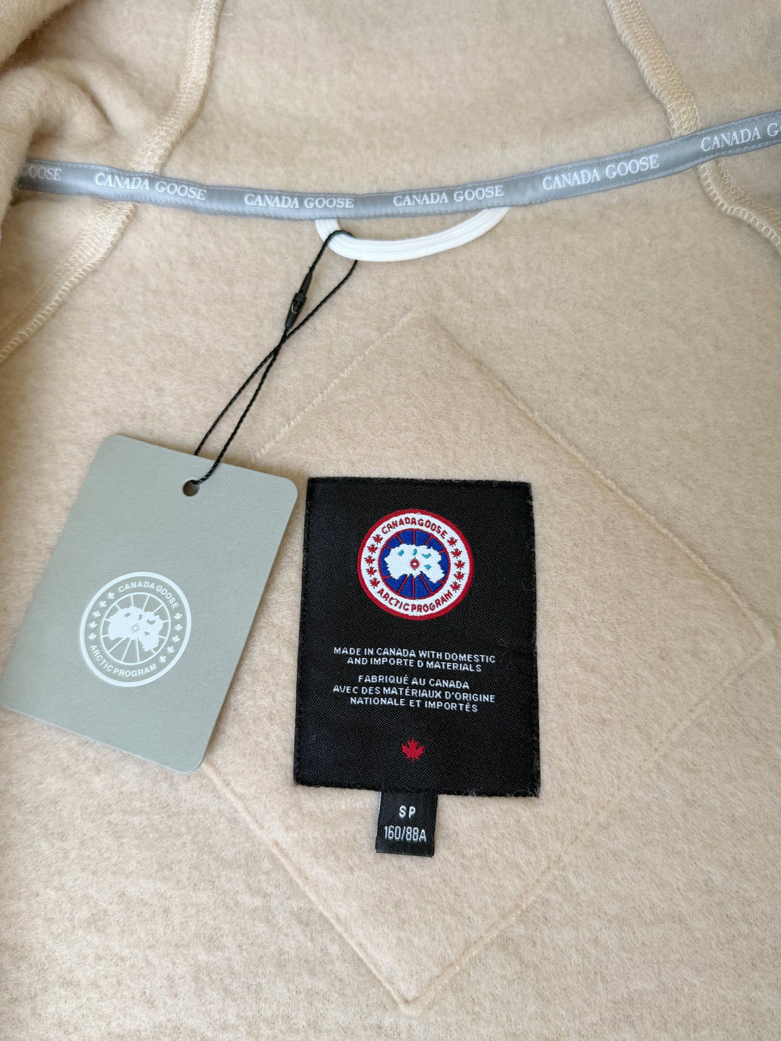 大鹅/Canada Goose抓绒羊毛饰章连帽夹克外套 面料选择: 以羊毛为主体，加入极少量氨纶和聚酯纤维混纺而成的羊毛抓绒面料。羊毛作为核心天然成分，提供无与伦比的温度调节、抑菌防臭和亲肤感。少量氨纶的喂入赋予了面料一定的四向弹性，日常活动时毫无束缚感。作为抓绒结构的基础，聚酯纤维增强了面料的耐久性、抗皱性和快干性，它构成了抓绒的蓬松感，并提供锁住空气等功能。 细节1: 衣袖和下摆采用2乘2罗纹收口，其强大的弹性，可以紧密贴合不同粗细的手腕和腰部，形成一个有效的&lsquo;密封&rsquo;,防止冷风倒灌。在多次拉伸和使用后仍能保持原有形状，不易松垮变形。且罗纹袖口能传递一种设计感，强化视觉美观度。 细节2: 兜帽设计与松紧扣的搭配提升穿戴时的保暖度与防风性能。 辅料:定制弹性抽绳松紧扣定制拉链吊坠定制臂章 尺码：S M L 颜色：黑 灰 咖 米 粉