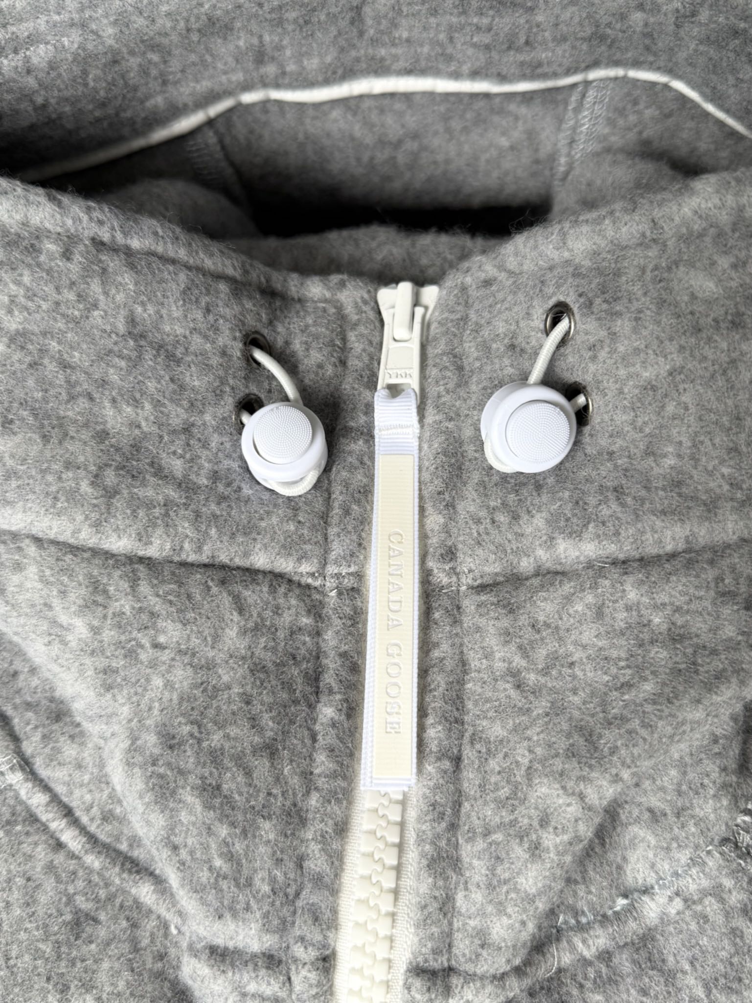 大鹅/Canada Goose抓绒羊毛饰章连帽夹克外套 面料选择: 以羊毛为主体，加入极少量氨纶和聚酯纤维混纺而成的羊毛抓绒面料。羊毛作为核心天然成分，提供无与伦比的温度调节、抑菌防臭和亲肤感。少量氨纶的喂入赋予了面料一定的四向弹性，日常活动时毫无束缚感。作为抓绒结构的基础，聚酯纤维增强了面料的耐久性、抗皱性和快干性，它构成了抓绒的蓬松感，并提供锁住空气等功能。 细节1: 衣袖和下摆采用2乘2罗纹收口，其强大的弹性，可以紧密贴合不同粗细的手腕和腰部，形成一个有效的&lsquo;密封&rsquo;,防止冷风倒灌。在多次拉伸和使用后仍能保持原有形状，不易松垮变形。且罗纹袖口能传递一种设计感，强化视觉美观度。 细节2: 兜帽设计与松紧扣的搭配提升穿戴时的保暖度与防风性能。 辅料:定制弹性抽绳松紧扣定制拉链吊坠定制臂章 尺码：S M L 颜色：黑 灰 咖 米 粉